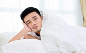 Man with black hair under the covers leaning on his hand, looking sad and sleepy.