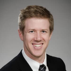 Matthew Carmody, DDS - Man smiling in a suit and tie against a grey background.