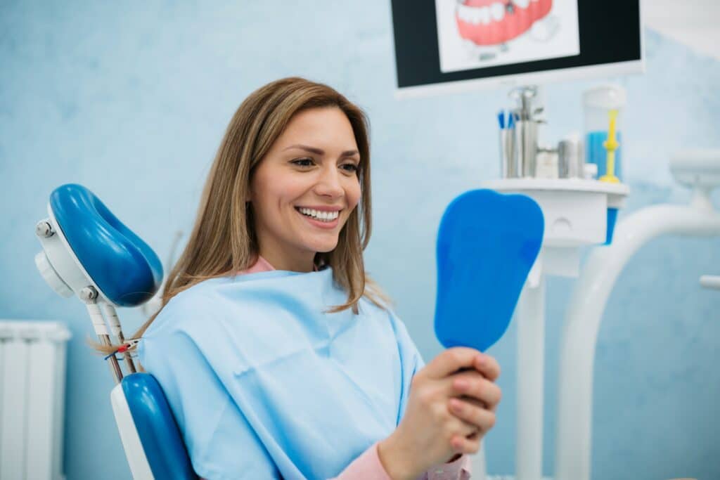 Young adult happy female patient relaxing while visiting a dentist office, holding a mirror and with a toothy smile on her face inspecting her teeth
