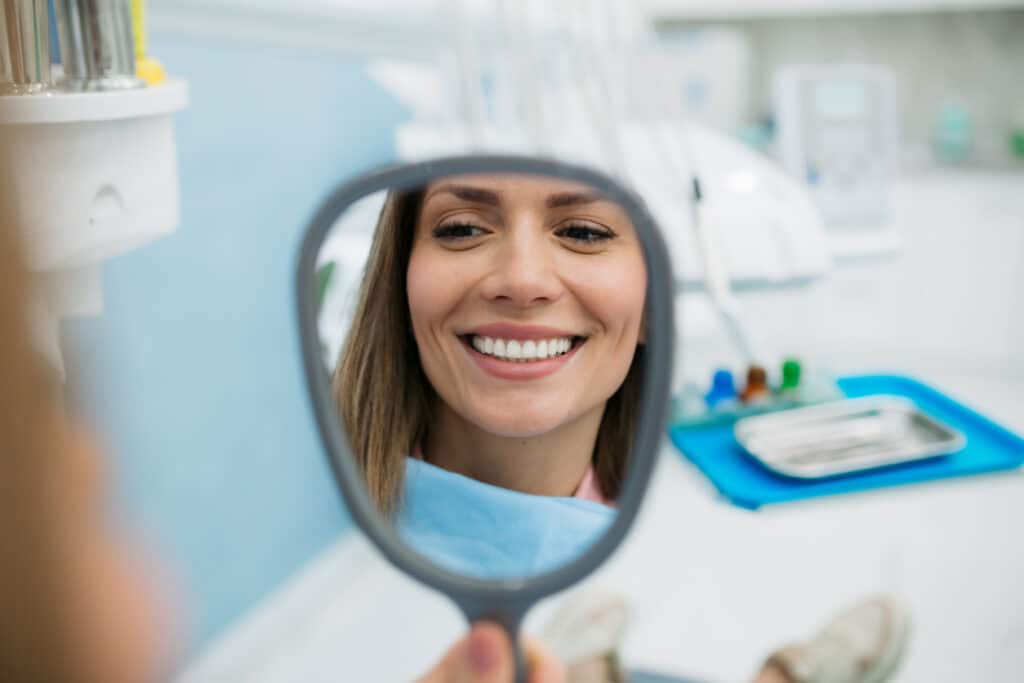 Young adult happy female patient relaxing in a chair while looking at her dental implants in a mirror.