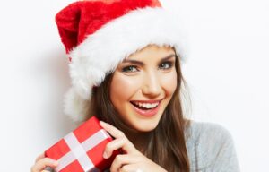 A woman in a Santa hat holding a present, smiling with clean white teeth
