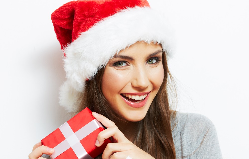 A woman in a Santa hat holding a present, smiling with clean white teeth
