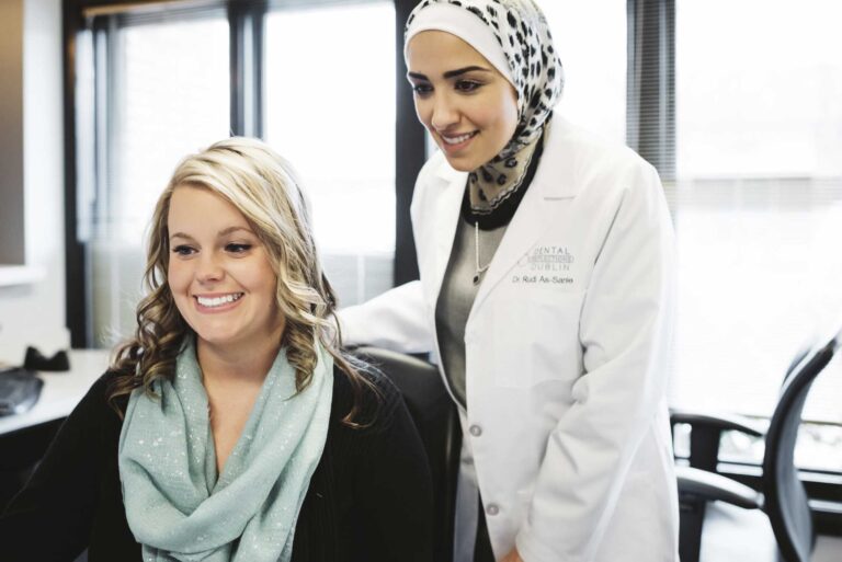 Two individuals in a professional setting, one seated with a light blue scarf, the other standing in a white lab coat labeled "DENTAL DUBLIN" and "Dr. Rudi As-Sanie," in front of large windows with blinds.