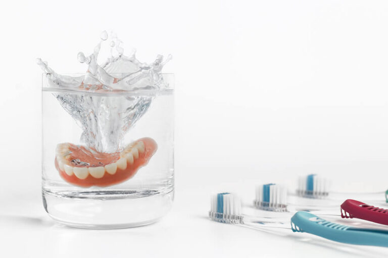 A set of dentures being dropped into a glass of water, creating a splash, with toothbrushes of different colors (blue, red, and green) next to the glass.