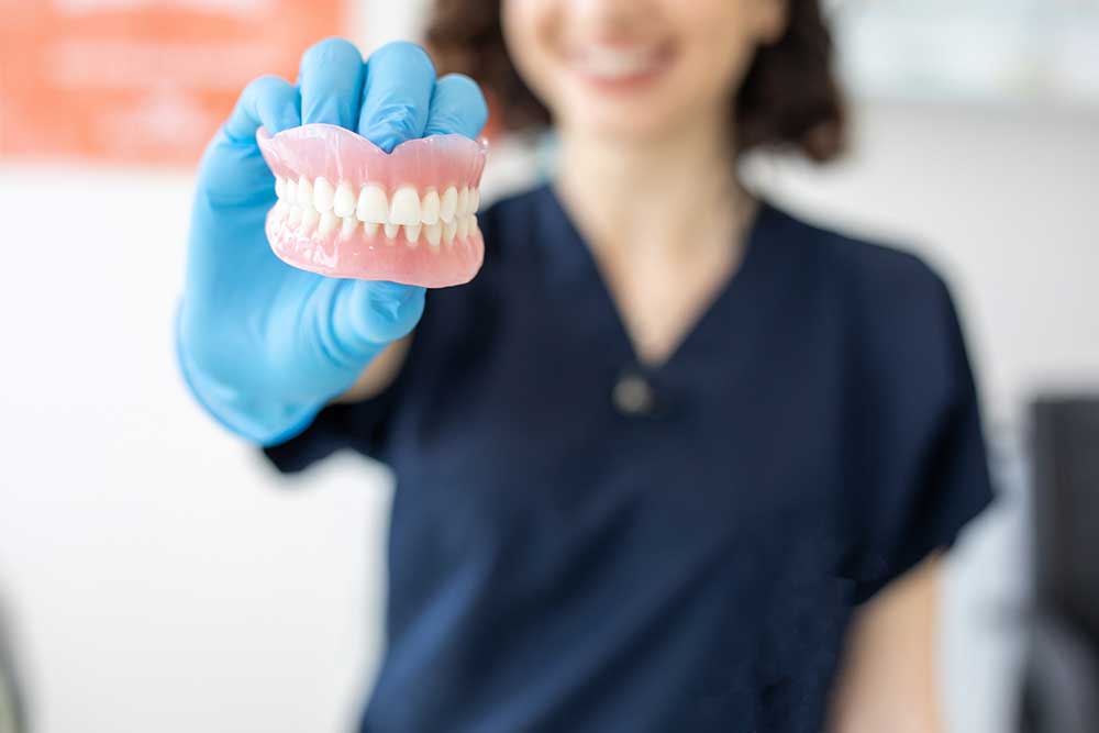 A dental professional in a dark blue uniform and blue gloves holding a set of dentures.