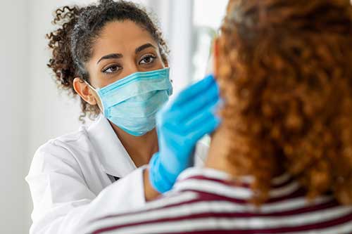 Healthcare professional in a white coat and blue gloves examining the neck of a person with curly hair and a striped shirt.
