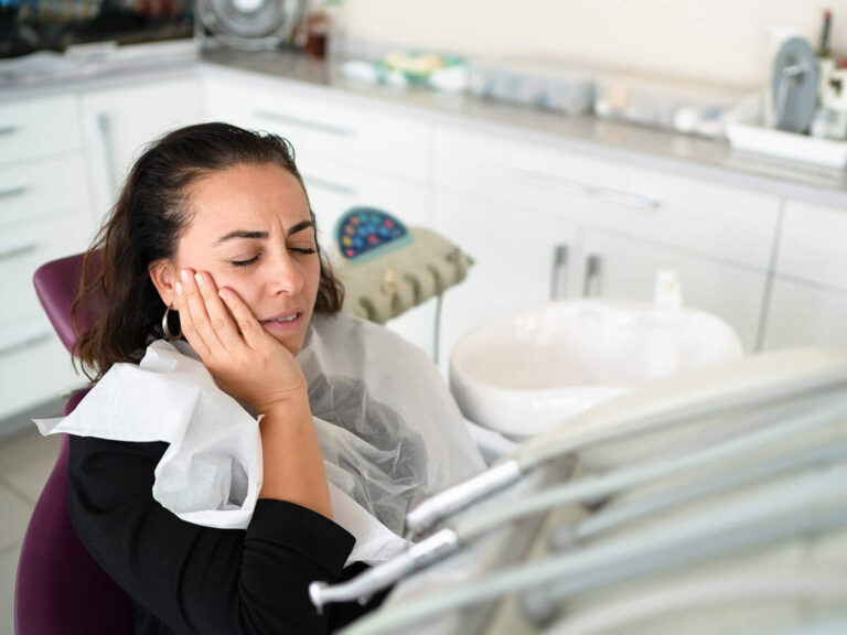 The image shows a woman sitting in a dental chair, covered with a protective bib, and holding her cheek, indicating pain. The background has dental equipment and tools, highlighting a dental clinic setting.