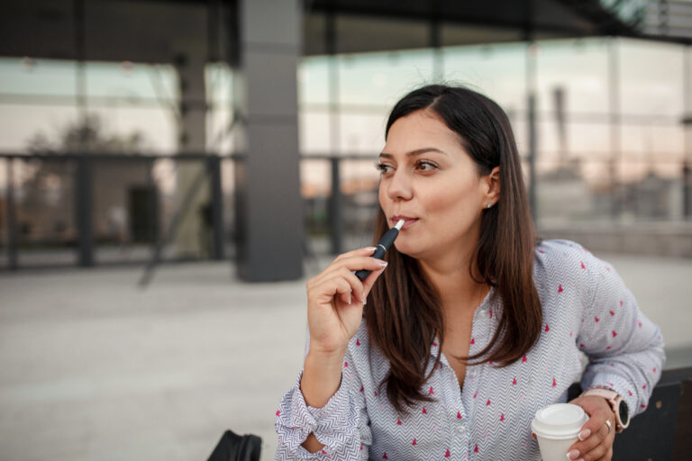 A person holding a white disposable cup with a lid in one hand and a cigarette in the other hand. They are wearing a light-colored shirt with a pattern of small red shapes. The background is an outdoor or semi-outdoor setting with glass windows and a modern architectural design.