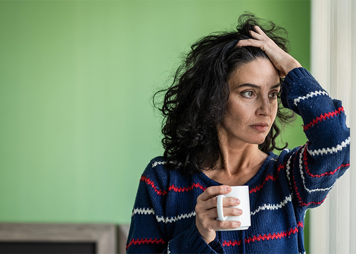 The image shows a person with curly hair wearing a blue sweater, holding a white mug in one hand and the other hand on their head, likely indicating stress.