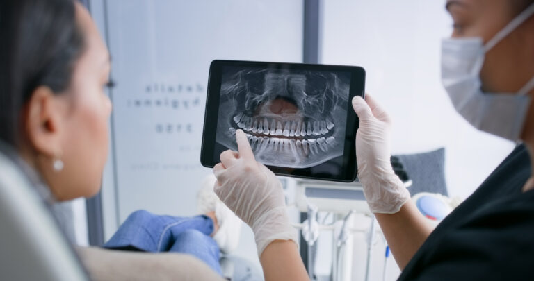A dental professional points at a specific area on a panoramic dental X-ray displayed on a tablet while a patient sits in a dental chair.