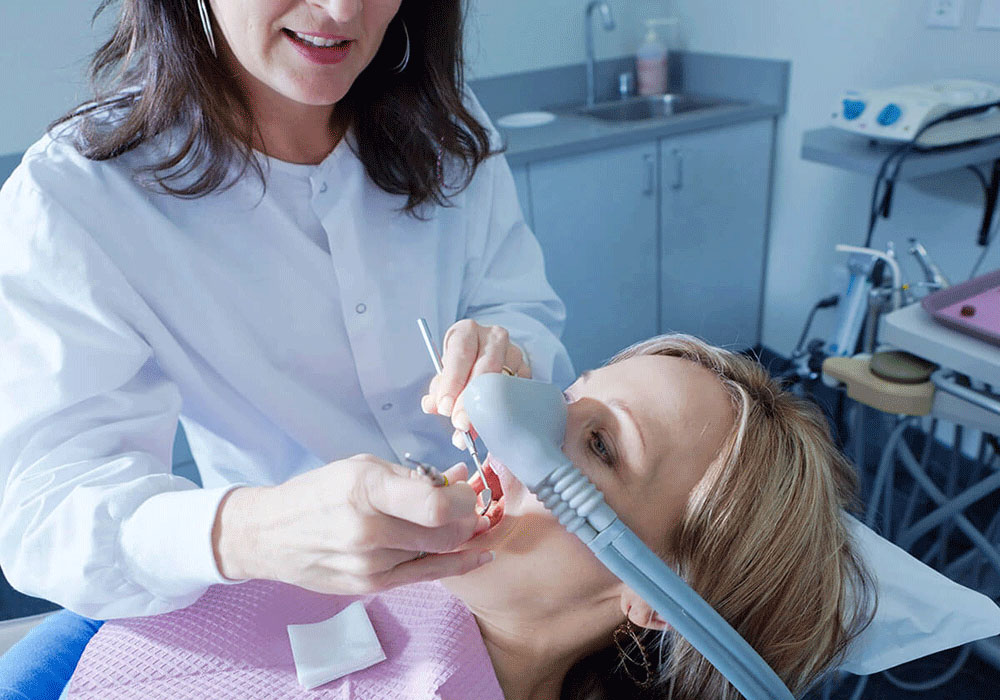 A dental professional performs a procedure on a patient in a dental chair, wearing a pink dental bib and a nasal mask, surrounded by dental office equipment.