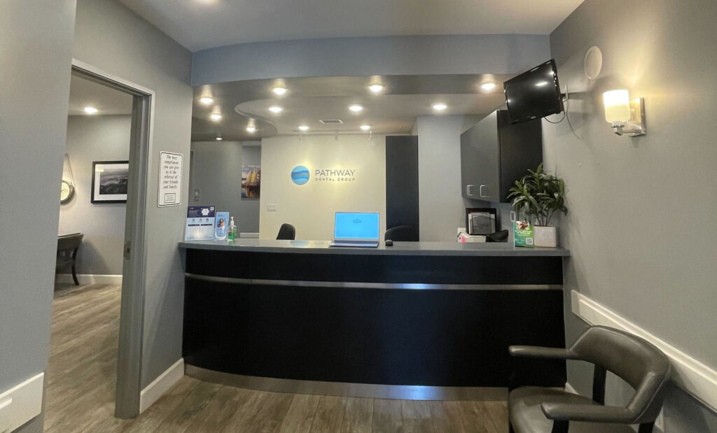 Reception area of a dental office named "Pathway Dental Group." Features a modern reception desk with a laptop and supplies, a logo on the wall, a small television, a plant, and modern decor. An open doorway leads to another room.