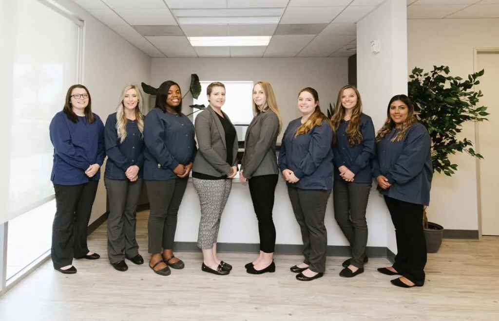 Eight professionally dressed individuals stand in an office, divided into two groups of four, facing each other. The modern space features light-colored flooring, walls, and two large plants, suggesting a corporate setting.