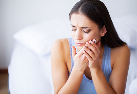 a woman holding her jaw in discomfort while sitting on a bed